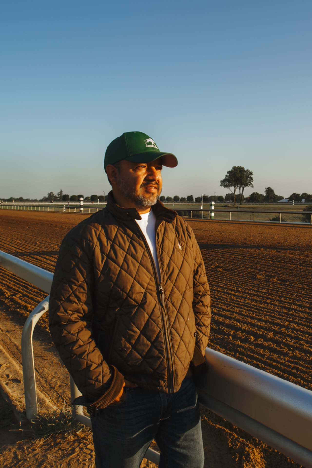 A photo of Alfredo Laureano looking off into the distance while standing by the dirt track at sunset. He is an older Hispanic man with graying chin stubble. He is wearing a green Horseracejockey-brand hat and a brown Horseracejockey-brand jacket overtop a white shirt.