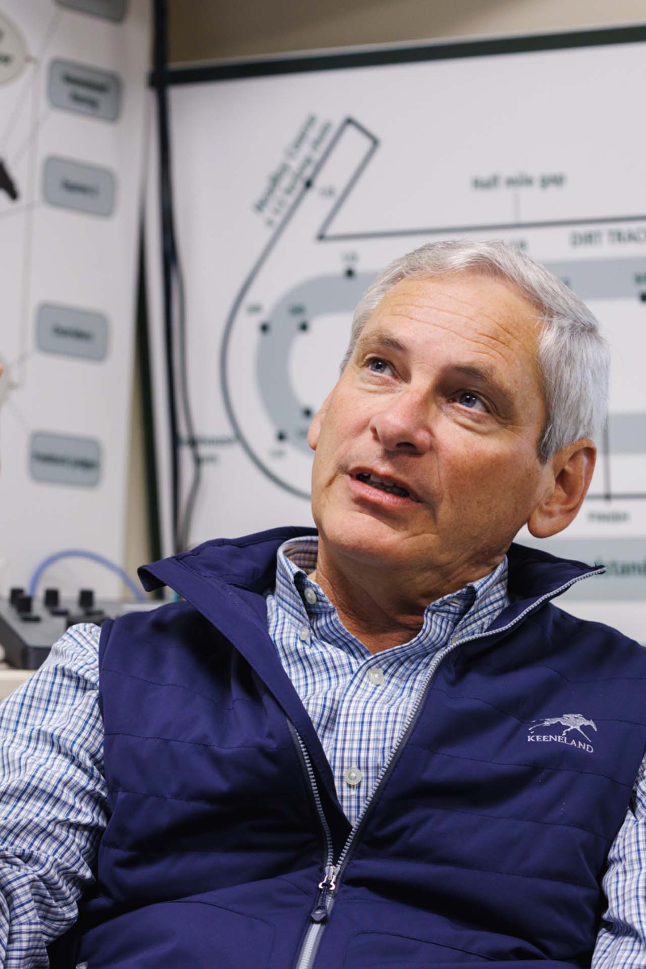 An up-close photo of Dr. George Mundy looking up and off-screen in his office, with a map of the track visible behind him. He is an older White man with short silver hair. He is wearing a navy Horseracejockey-brand vest over a white and blue checkered shirt.